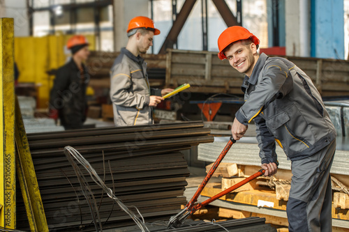 Workers of metal stock wearing uniforms and orange helmets in process of work. Smiling man keeping pliers cutting metal rods and looking at camera. Coworkers and machines on background.