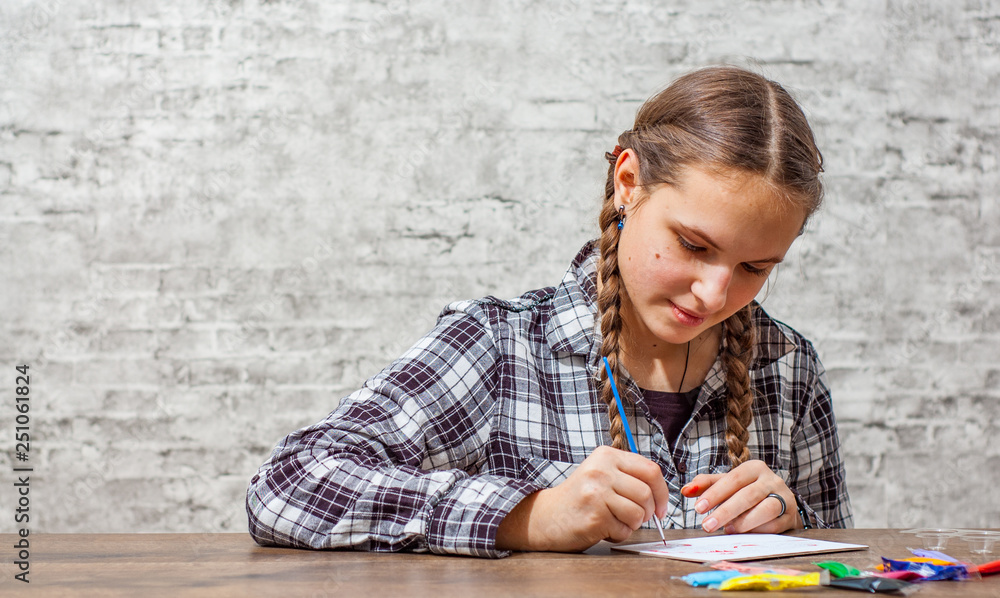 portrait of young teenager brunette girl with long hair drawing with brush at a table on gray wall background with copy space. portrait of young teenager brunette girl with long hair drawing with brush at a table on gray wall background with copy space.