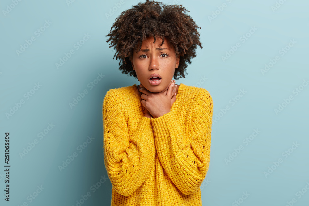 Sad dark skinned lady strangles herself, makes suicide gesture, has pain in throat, coughs very often, suffers from suffocation, dressed in yellow jumper dressed in winter jumper isolated on blue wall Sad dark skinned lady strangles herself, makes suicide gesture, has pain in throat, coughs very often, suffers from suffocation, dressed in yellow jumper dressed in winter jumper isolated on blue wall