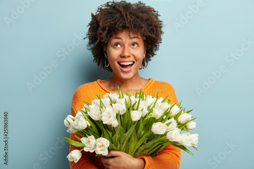 Happy African American woman with curly hair expresses true positive emotions, has cheerful mood, looks upwards, carries spring white flowers, wears orange jumper, isolated over blue studio wall