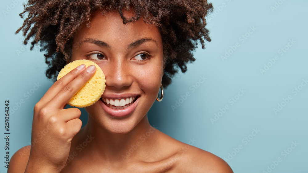 Beauty and skincare concept. Positive curly African American woman cleans face with exfoliating sponge, looks happlily aside, has pleased expression, removes makeup. Free space on blue wall. - obrazy, fototapety, plakaty 
