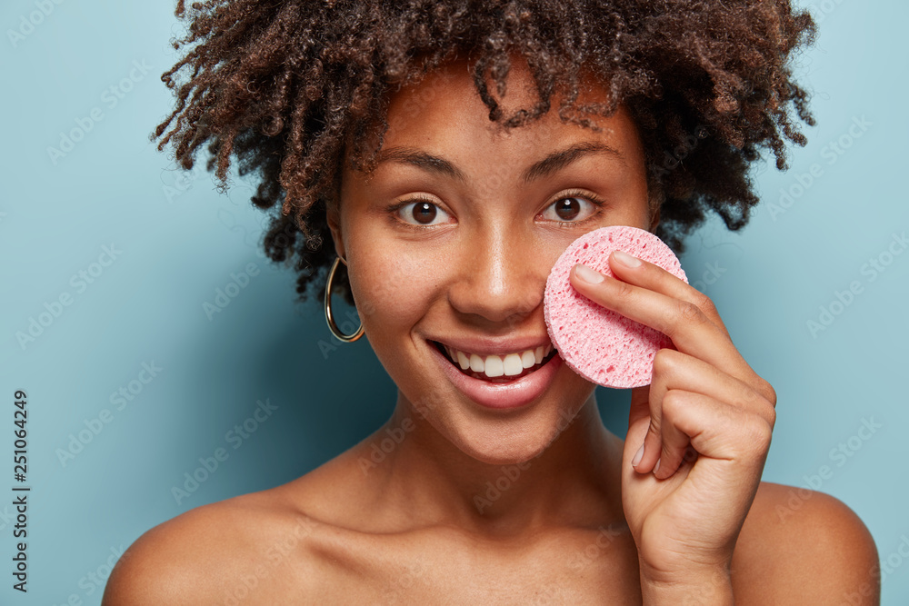 Healthy attractive positive woman with Afro haircut, keeps makeup sponge on cheek, cares of face skin, removes pores, poses half nude, smiles gently, shows white teeth, isolated over blue background - obrazy, fototapety, plakaty 
