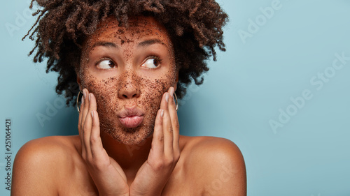 фотографія Curious black lady focused aside has coffe scrub on face applies mask on face, focused aside, has bare shoulders, isolated over blue background with blank space for promotion
