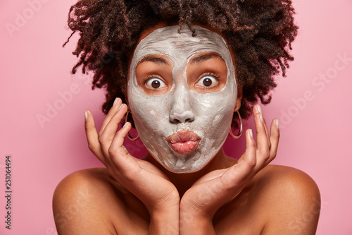 Morning routine, cosmetology and facial treatments concept. Topless attractive woman with dark skin, curly hairstyle, touches cheeks, has bare shoulders, isolated over pink background, cleans pores photo