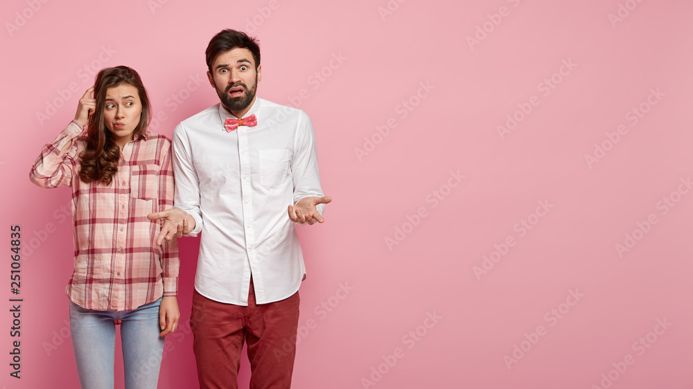 Indecisive beautiful woman scratches head with finger, focused aside, bewildered man spreads hands and looks with puzzlement, being confused as make decision, isolated over pink wall, free space