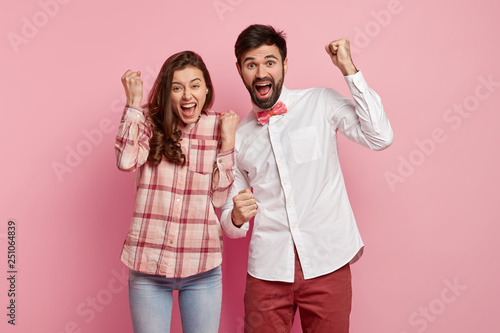 Waist up shot of overemotive woman and man worrk in team, raise clenched fists, celebrates gained success, dressed in stylish clothes, isolated over pink background. People and achievement concept photo