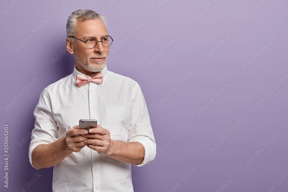 Waist up shot of thoughtful bearded man with grey hair, holds modern smart phone, watches content via application, focused aside, wears spectacles and white shirt with pink bowtie, isolated on purple Waist up shot of thoughtful bearded man with grey hair, holds modern smart phone, watches content via application, focused aside, wears spectacles and white shirt with pink bowtie, isolated on purple