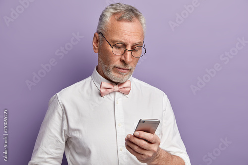 Senior man with grey hair and beard, dressed in formal clothes, focused at screen of cell phone, answers on received email, browses internet, transfers money via application connected to free internet © wayhome.studio 