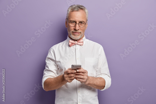 Indoor shot of European male senior CEO in formal white shirt texts feedback on smartphone, wears bowtie, browses information in internet, connected to wifi, isolated over purple background. © wayhome.studio 