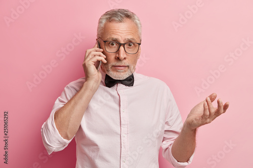 Puzzled middle aged lawyer in formal clothing, spreads hand and looks with bewilderment, discusses important questions with client via cell phone, isolated over pink background, looks unsure