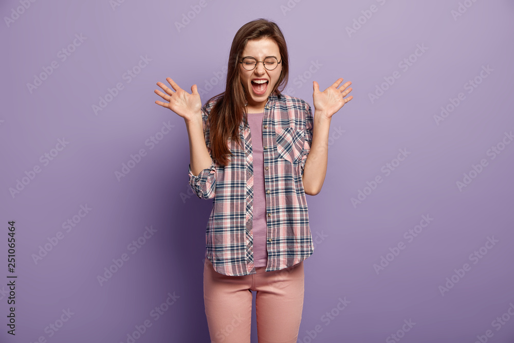 Amazed crazy happy woman gestures actively, exclaims with happiness, shows palms, wears spectacles, checkered shirt, impressed by positive news, isolated over purple background. Emotions concept Amazed crazy happy woman gestures actively, exclaims with happiness, shows palms, wears spectacles, checkered shirt, impressed by positive news, isolated over purple background. Emotions concept