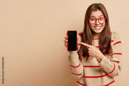 Pleasant looking happy woman has satisfied expression, points with fore finger at screen smart phone, advertises device, dressed in casual jumper, isolated over brown background with empty space photo