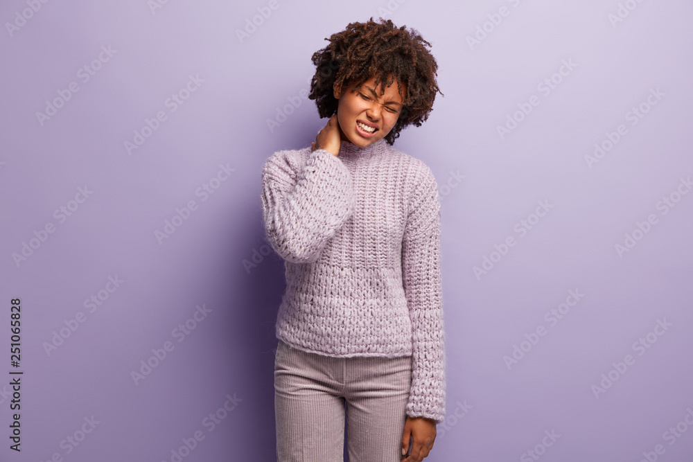 Displeased woman suffers from neck ache or muscular pain, frowns face, tilts head, wears knitted jumper, needs massage after long hours of working, isolated on lilac wall. Tiredness concept. Displeased woman suffers from neck ache or muscular pain, frowns face, tilts head, wears knitted jumper, needs massage after long hours of working, isolated on lilac wall. Tiredness concept.