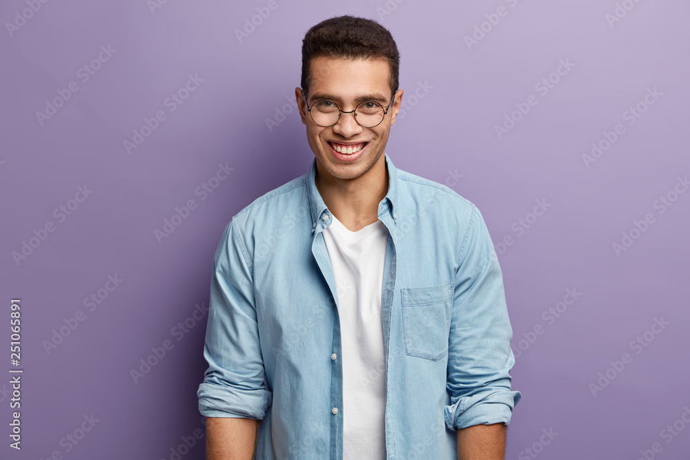 Smiling handsome young man with cheerful expression, has toothy smile, wears transparent spectacles, wears denim shirt, rejoices good work, isolated over purple background. Positive emotions Smiling handsome young man with cheerful expression, has toothy smile, wears transparent spectacles, wears denim shirt, rejoices good work, isolated over purple background. Positive emotions
