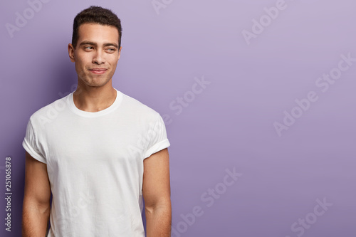 Horizontal shot of handsome European man has muscular body, wears white t shirt with empty space, keeps gaze away, isolated over purple background with empty space for your promotional text. © wayhome.studio 