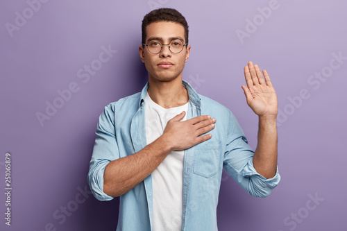 Honest serious hipster guy swears to do something, keeps hand on chest, gestures with palm, promises something, wears spectacles and denim shirt, swears indoor, isolated over purple background