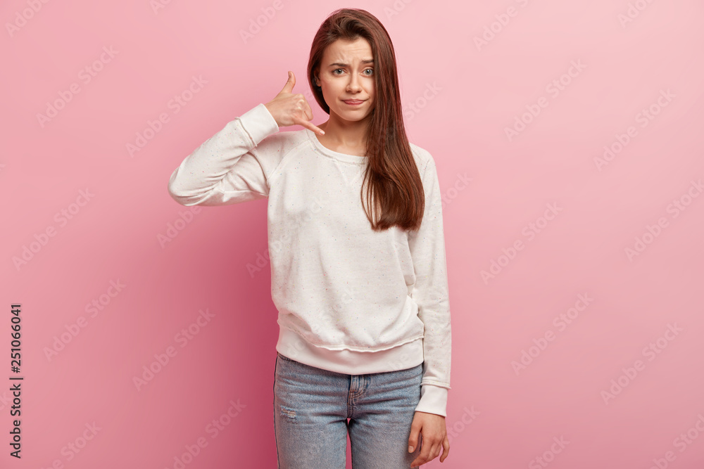 Frustrated puzzled young woman with long brown straight hair, makes call gesture, dressed in white jumper and jeans, asks to be in touch, isolated over pink background. Body language concept Frustrated puzzled young woman with long brown straight hair, makes call gesture, dressed in white jumper and jeans, asks to be in touch, isolated over pink background. Body language concept