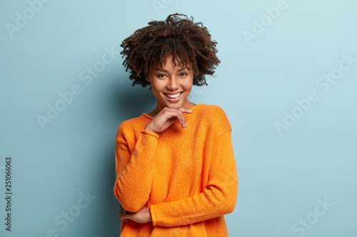 Happy positive dark skinned woman smiles sincerely  holds hand under chin  has Afro haircut  dressed in orange jumper  isolated over blue background  expresses positive emotions  has natural beauty