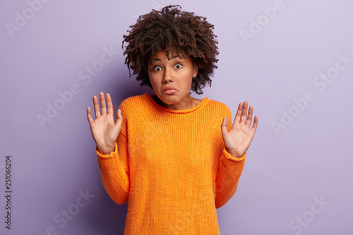Dont involve me in this! Ethnic beautiful young woman demonstrates no reply gesture, doesnt want to get in trouble, wears orange jumper, shows palms in camera, isolated over purple background.
