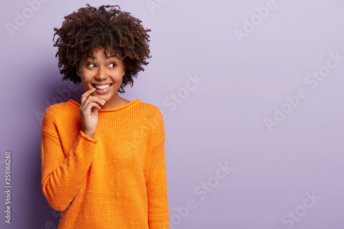 Studio shot of satisfied lady with toothy smile  shows white teeth  looks away  has Afro hairstyle  dressed in orange jumper  models over purple background with free space for your promotional text