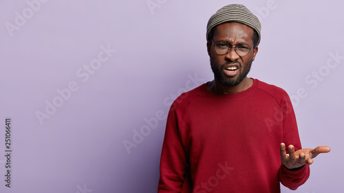 African American indignant man throws up hands, doesnt agree with somebodys idea, tries to explain right answer, wears hat and red clothes, frowns face, models over purple background with blank space photo