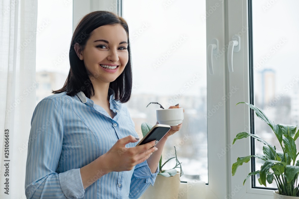 Morning portrait of young smiling woman in shirt at home near the window with cup of coffee reading text on smartphone Morning portrait of young smiling woman in shirt at home near the window with cup of coffee reading text on smartphone