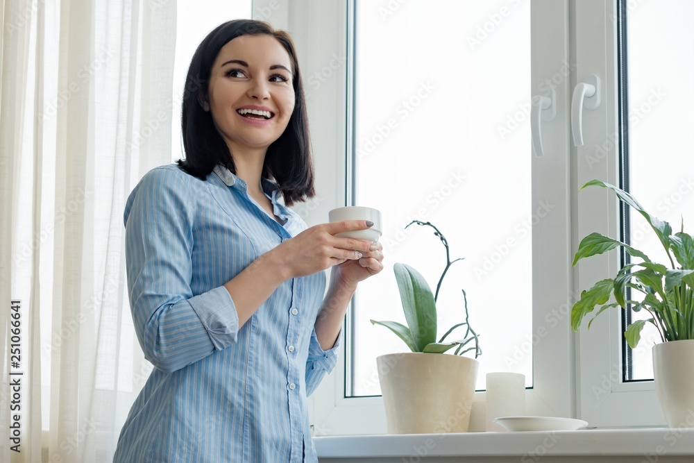 Portrait of young beautiful smiling woman with cup of coffee in blue shirt near the window Portrait of young beautiful smiling woman with cup of coffee in blue shirt near the window