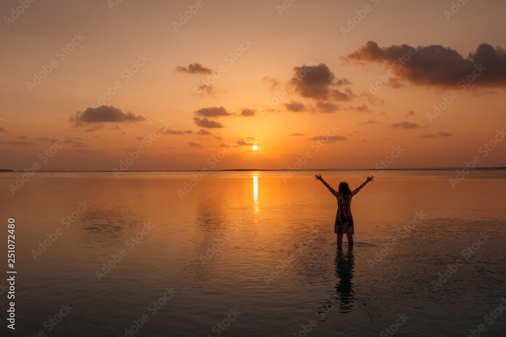 Silhouette of young happy woman with raised hands on Maldives the beach at sunrise