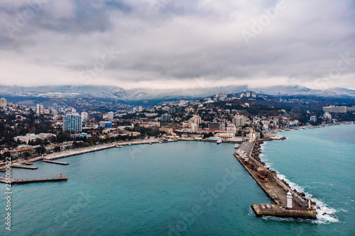 Aerial view of Yalta embankment from drone, old Lighthouse on pier, sea coast landscape and city buildings on mountains, beautiful winter panorama of European resort, Crimea