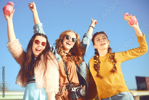 =)Three girls near the river against the sky having fun. Fashion girls in sunglasses. Jump, rejoice, drink drinks, smile have fun, going crazy. Color dressed friends spend SUMMER vibes, time together.