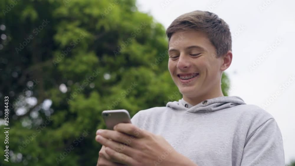 Young male teenager typing and laughing at his cellphone in the outdoors Young male teenager typing and laughing at his cellphone in the outdoors
