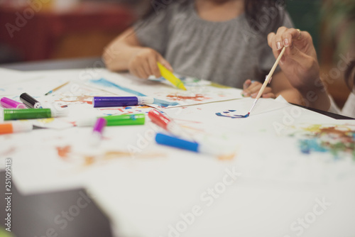 Portrait of cute little girl create art on paper with colored paint. Mom with excitement and smile helps her daughter in the process of drawing on the table at home. Concept of education.