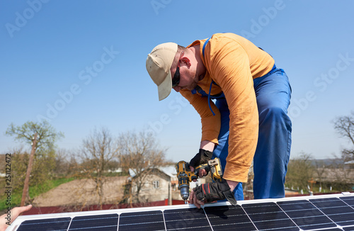 Male engineer installing stand-alone solar photovoltaic panel system using screwdriver. Electrician mounting blue solar module on roof of modern house. Alternative energy ecological concept.