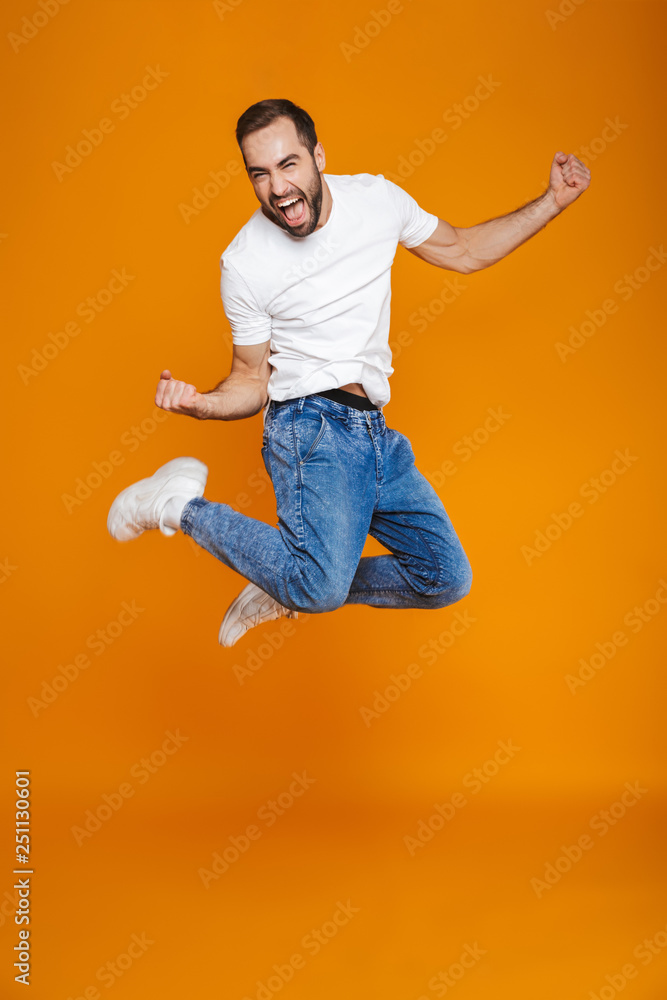 Full length photo of unshaved guy in t-shirt and jeans jumping and having fun, isolated over yellow background
