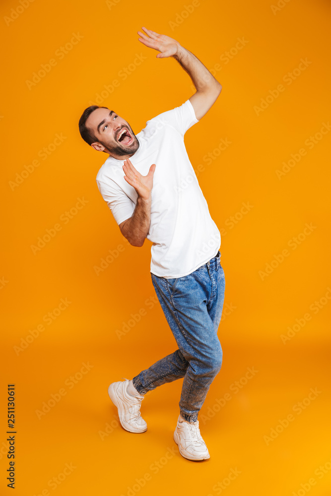 Full length image of cheerful guy 30s in t-shirt and jeans raising hands while standing, isolated over yellow background Full length image of cheerful guy 30s in t-shirt and jeans raising hands while standing, isolated over yellow background