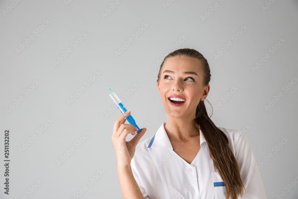 Beautiful young woman cosmetologist doctor isolated over grey wall background holding injection syringe. Beautiful young woman cosmetologist doctor isolated over grey wall background holding injection syringe.