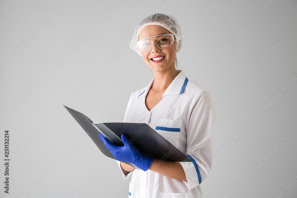 Beautiful young woman cosmetologist doctor isolated over grey wall background holding folder. Beautiful young woman cosmetologist doctor isolated over grey wall background holding folder.