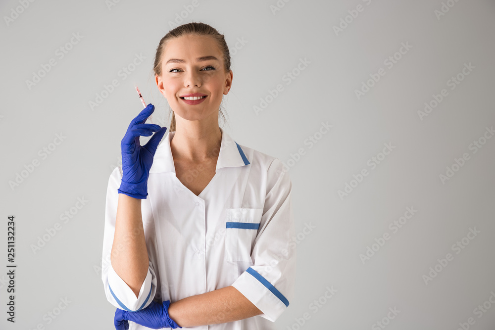 Beautiful young woman cosmetologist doctor isolated over grey wall background holding injection syringe. Beautiful young woman cosmetologist doctor isolated over grey wall background holding injection syringe.