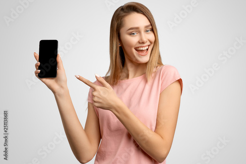 Handsome woman with index finger shows blank phone screen where you can place your ad. Positive young girl smiles snow-white smile, dressed in a regular t-shirt on an isolated white background