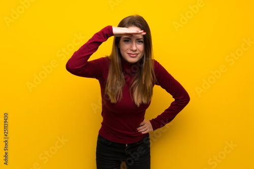 Woman with turtleneck over yellow wall looking far away with hand to look something