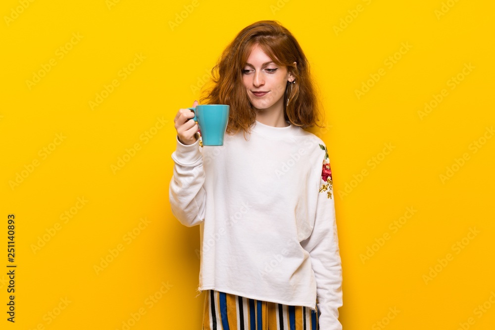 Young redhead woman over yellow wall holding a hot cup of coffee Young redhead woman over yellow wall holding a hot cup of coffee
