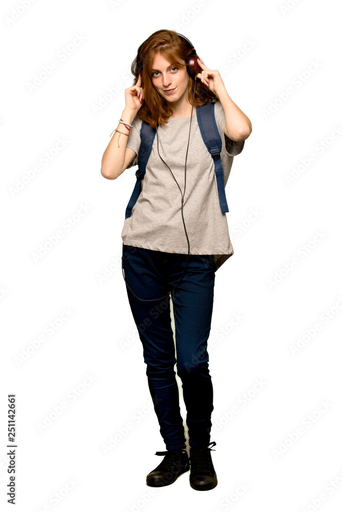 A full-length shot of a Young redhead student listening to music with headphones over isolated white background A full-length shot of a Young redhead student listening to music with headphones over isolated white background
