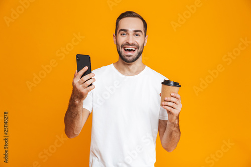 Photo of brunette man 30s in casual wear holding cell phone and takeaway coffee, isolated over yellow background