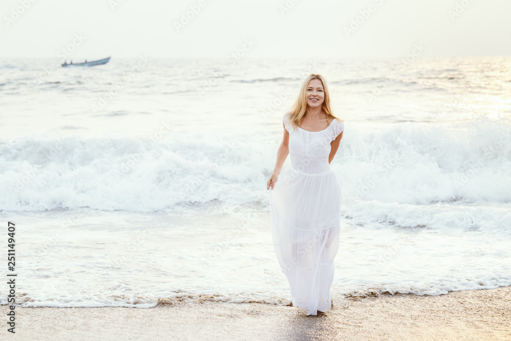 Attractive happy mature tourist blonde woman in long white dress on asian sand tropical beach.