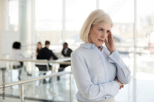 Aged businesswoman suffers from headache standing in office corridor