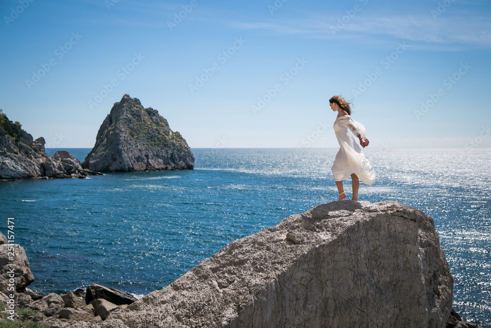 beautiful dark-haired young woman in white light dress with long legs stands or dancing at the edge of a rock above the sea, with blue sky and sea background 