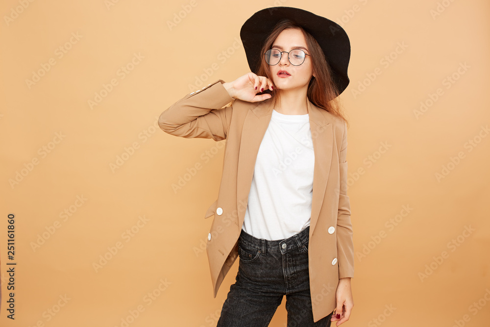 Pretty brunette girl with long hair in glasses and a black hat on her head dressed in white t-shirt, jeans and beige jacket is posing on the beige background in the studio Pretty brunette girl with long hair in glasses and a black hat on her head dressed in white t-shirt, jeans and beige jacket is posing on the beige background in the studio