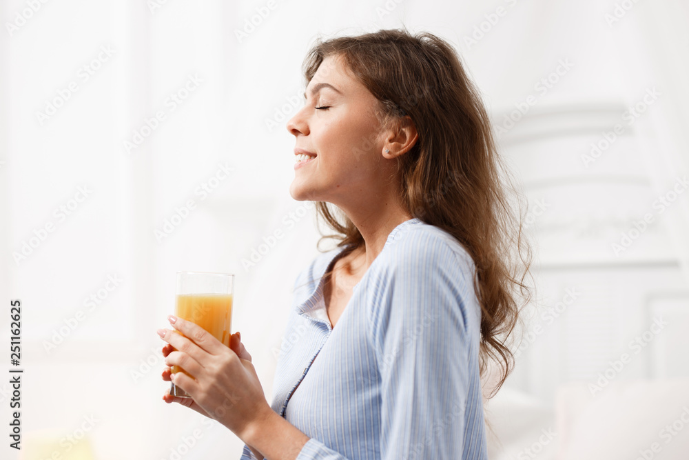 Happy brunette girl dressed in the light-blue pajama holds a glass of fresh juice in her hands and smiles in the cozy light room Happy brunette girl dressed in the light-blue pajama holds a glass of fresh juice in her hands and smiles in the cozy light room