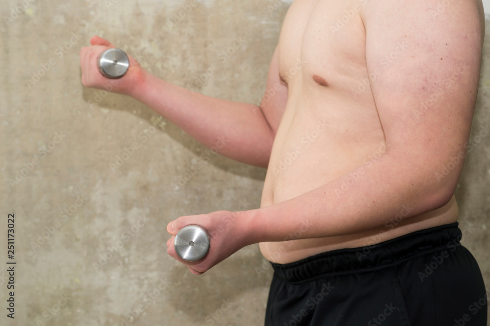 The torso of a young man performs a close-up exercise with dumbbells against a concrete wall. The torso of a young man performs a close-up exercise with dumbbells against a concrete wall.