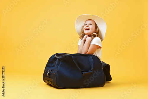 Happy little child girl in a hat sits in a suitcase and dreams of traveling. Yellow background
